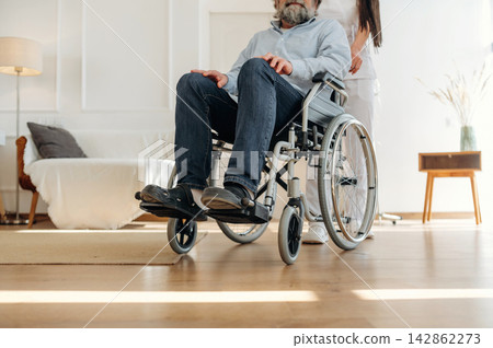 Elderly man in wheelchair being assisted by caregiver in a bright living room with wooden flooring and modern furniture, creating a supportive environment Elderly man in wheelchair being assisted by caregiver in a bright living room with wooden flooring and modern furniture, creating a supportive environment 142862273