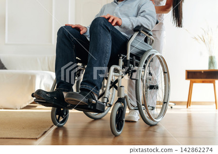 Male wheelchair user in casual clothing being assisted by caregiver in a bright indoor living space with a sofa and wooden table visible in the background Male wheelchair user in casual clothing being assisted by caregiver in a bright indoor living space with a sofa and wooden table visible in the background 142862274