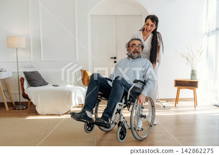 Woman assisting elderly man in wheelchair inside a bright living room with modern decor, featuring a sofa, lamp, and wooden furniture in the background Woman assisting elderly man in wheelchair inside a bright living room with modern decor, featuring a sofa, lamp, and wooden furniture in the background 142862275