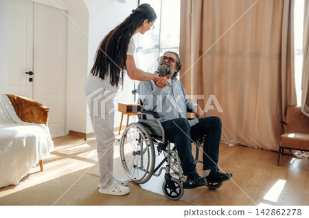 Female caregiver in white uniform assists elderly man in wheelchair during home visit, bright interior with natural light and soft furnishings visible in the background Female caregiver in white uniform assists elderly man in wheelchair during home visit, bright interior with natural light and soft furnishings visible in the background 142862278