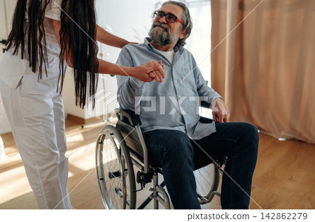 Elderly man in wheelchair receiving assistance from caregiver in white uniform inside a well-lit room with wooden flooring and soft curtains 142862279