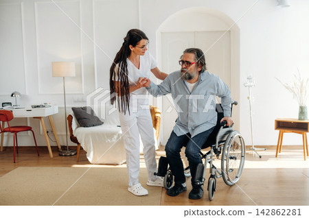 Female caregiver assisting elderly man in wheelchair to stand inside a modern living room with wooden flooring and minimalistic furniture Female caregiver assisting elderly man in wheelchair to stand inside a modern living room with wooden flooring and minimalistic furniture 142862281