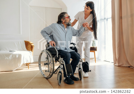 Male wheelchair user receives assistance from a female caregiver in a bright, modern living room with large windows and wooden flooring Male wheelchair user receives assistance from a female caregiver in a bright, modern living room with large windows and wooden flooring 142862282