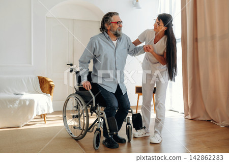 Middle-aged man in wheelchair assisted by female caregiver in white uniform inside a well-lit room with soft furnishings and natural light streaming through the window 142862283
