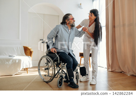 Middle-aged man in wheelchair receives assistance from female caregiver in modern living room with natural light and soft furnishings Middle-aged man in wheelchair receives assistance from female caregiver in modern living room with natural light and soft furnishings 142862284