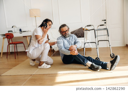 Female caregiver in white scrubs assists elderly man sitting on floor with walker nearby, modern interior with wooden flooring and minimalistic furniture visible in the background 142862290