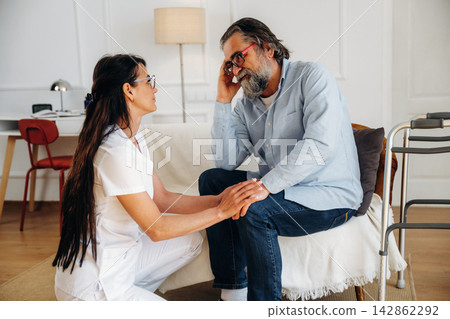 Female caregiver in white uniform attentively listens to elderly man in casual attire sitting on a couch in a well-lit living room with modern decor 142862292