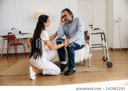 Female caregiver in white uniform assists elderly man with gray hair sitting on chair in modern home interior with walker and desk in the background Female caregiver in white uniform assists elderly man with gray hair sitting on chair in modern home interior with walker and desk in the background 142862293