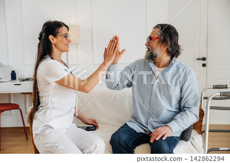 Female healthcare professional in white uniform interacts with elderly male patient in casual attire, both smiling and engaging in a friendly hand gesture indoors 142862294