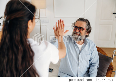 Elderly man with gray beard and glasses smiling while giving a high-five to a young woman with long hair in a bright indoor living room setting Elderly man with gray beard and glasses smiling while giving a high-five to a young woman with long hair in a bright indoor living room setting 142862295