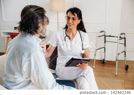 Female healthcare professional in white scrubs discusses treatment with male patient sitting on a couch in a well-lit room with a walker nearby Female healthcare professional in white scrubs discusses treatment with male patient sitting on a couch in a well-lit room with a walker nearby 142862296