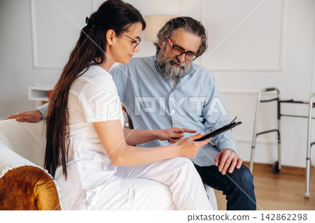 Female healthcare professional in white uniform discusses treatment plan with elderly male patient in a bright, modern living room setting with mobility aid in background Female healthcare professional in white uniform discusses treatment plan with elderly male patient in a bright, modern living room setting with mobility aid in background 142862298