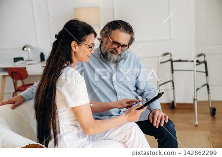 Female healthcare professional in white uniform discusses treatment options with elderly male patient in a well-lit room with modern decor and mobility aid nearby Female healthcare professional in white uniform discusses treatment options with elderly male patient in a well-lit room with modern decor and mobility aid nearby 142862299