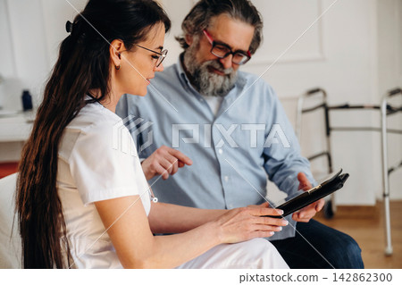 Female healthcare professional in white uniform discusses medical information with senior man in glasses, seated in a bright consultation room with medical equipment visible 142862300