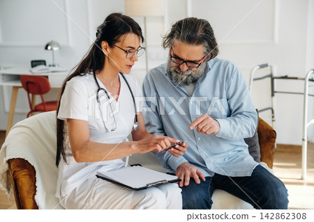 Female healthcare professional in white scrubs discusses treatment options with senior man in casual attire, sitting in a well-lit room with medical equipment visible Female healthcare professional in white scrubs discusses treatment options with senior man in casual attire, sitting in a well-lit room with medical equipment visible 142862308