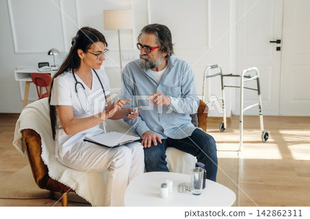 Female healthcare professional in white scrubs discusses medication with senior male patient in a well-lit living room with modern furniture and a walker nearby Female healthcare professional in white scrubs discusses medication with senior male patient in a well-lit living room with modern furniture and a walker nearby 142862311