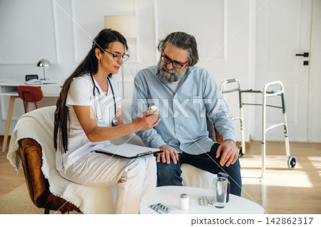 Female healthcare professional in white scrubs discusses medication with elderly male patient in a bright, modern living room with a walker and medical supplies Female healthcare professional in white scrubs discusses medication with elderly male patient in a bright, modern living room with a walker and medical supplies 142862317
