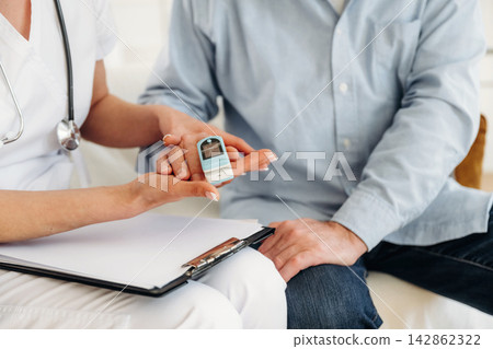 Healthcare professional in white scrubs demonstrates a blood glucose meter to a male patient seated on a couch, clipboard and medical documents visible on the side Healthcare professional in white scrubs demonstrates a blood glucose meter to a male patient seated on a couch, clipboard and medical documents visible on the side 142862322
