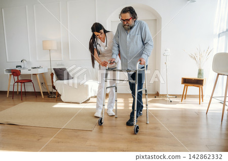 Female healthcare professional assists elderly man using walker in bright, modern living space with wooden flooring and minimalistic furniture Female healthcare professional assists elderly man using walker in bright, modern living space with wooden flooring and minimalistic furniture 142862332