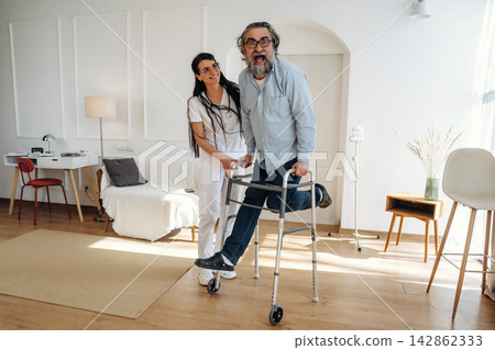 Female healthcare worker assists elderly man using walker in modern living room with bright lighting, comfortable furniture, and minimalistic decor Female healthcare worker assists elderly man using walker in modern living room with bright lighting, comfortable furniture, and minimalistic decor 142862333
