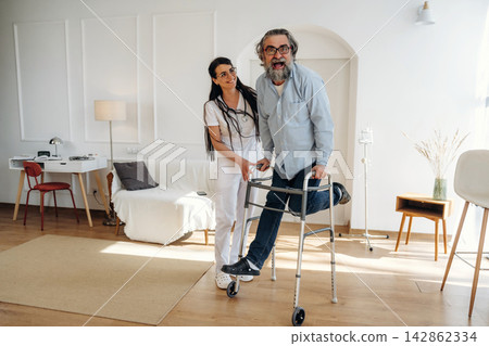 Female healthcare worker assists elderly man using walker in bright, modern living room with wooden floor and minimalistic furniture, creating a supportive environment for mobility Female healthcare worker assists elderly man using walker in bright, modern living room with wooden floor and minimalistic furniture, creating a supportive environment for mobility 142862334
