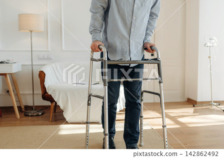 Male individual using a walker for support while standing in a bright, modern living room with a sofa and lamp in the background 142862492