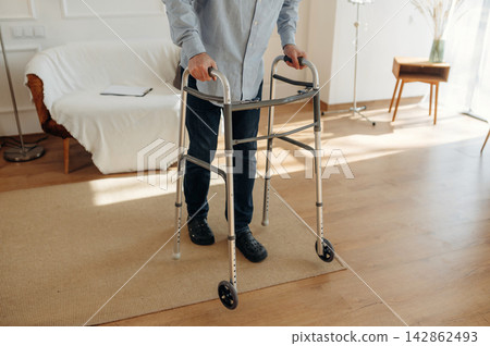 Elderly man using a walker for support while standing on a wooden floor in a bright, modern living room with a sofa and decorative elements Elderly man using a walker for support while standing on a wooden floor in a bright, modern living room with a sofa and decorative elements 142862493