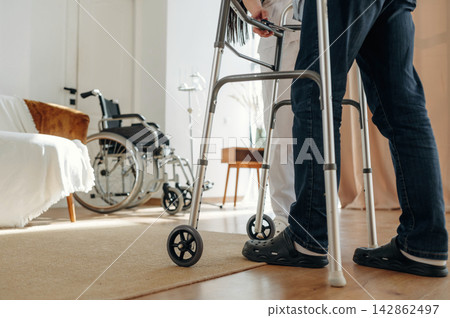 Person using a walker for support while assisted by a caregiver in a well-lit indoor setting with a wheelchair and furniture visible in the background Person using a walker for support while assisted by a caregiver in a well-lit indoor setting with a wheelchair and furniture visible in the background 142862497
