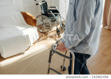 Elderly man using a walker in a well-lit living room with a wheelchair and a sofa covered in white fabric, showcasing a comfortable home environment 142862498
