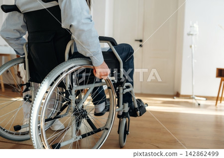 Individual in wheelchair navigating a well-lit room with wooden flooring, showcasing a comfortable and accessible living space with a door and medical equipment in the background Individual in wheelchair navigating a well-lit room with wooden flooring, showcasing a comfortable and accessible living space with a door and medical equipment in the background 142862499