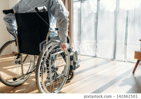 Individual in a wheelchair navigating a sunlit room with large windows and sheer curtains, showcasing a modern interior design and wooden flooring Individual in a wheelchair navigating a sunlit room with large windows and sheer curtains, showcasing a modern interior design and wooden flooring 142862501