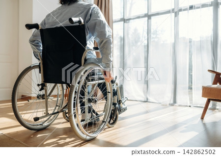 Individual in wheelchair facing large windows with sheer curtains, sunlight illuminating wooden floor and modern interior design elements in a spacious room Individual in wheelchair facing large windows with sheer curtains, sunlight illuminating wooden floor and modern interior design elements in a spacious room 142862502