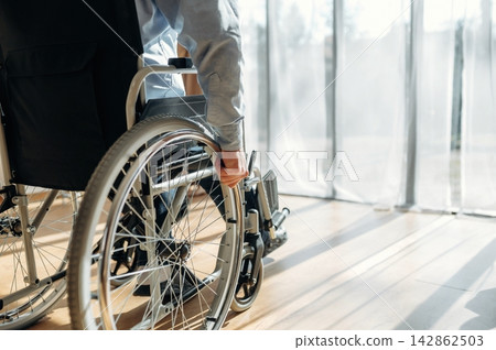 Individual in a wheelchair with gray clothing grips the wheel while positioned near a large window with sheer curtains and wooden flooring in a well-lit interior space Individual in a wheelchair with gray clothing grips the wheel while positioned near a large window with sheer curtains and wooden flooring in a well-lit interior space 142862503
