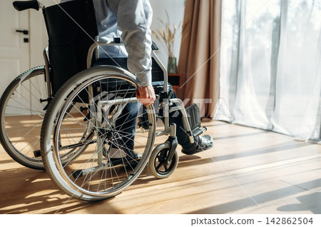 Individual in a wheelchair maneuvering on wooden floor near large windows with sheer curtains, showcasing a bright and airy indoor environment Individual in a wheelchair maneuvering on wooden floor near large windows with sheer curtains, showcasing a bright and airy indoor environment 142862504