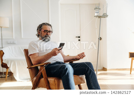 Mature man with gray beard sitting in wooden chair using smartphone while charging in bright living room with modern decor and natural light 142862527