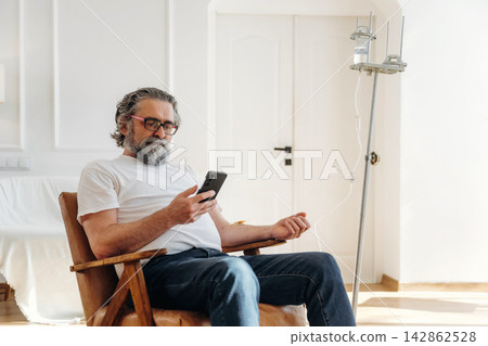 Senior man with gray hair and beard sitting in wooden chair using smartphone while receiving IV treatment in bright, modern room with white walls and natural light 142862528
