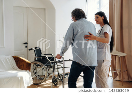 Healthcare professional assists elderly man using walker in a well-lit room with a wheelchair and comfortable seating area visible in the background Healthcare professional assists elderly man using walker in a well-lit room with a wheelchair and comfortable seating area visible in the background 142863059