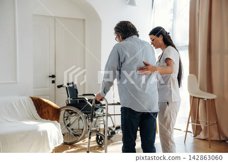 Caregiver assisting elderly man with walker in bright, modern living room featuring wheelchair and comfortable seating arrangements Caregiver assisting elderly man with walker in bright, modern living room featuring wheelchair and comfortable seating arrangements 142863060