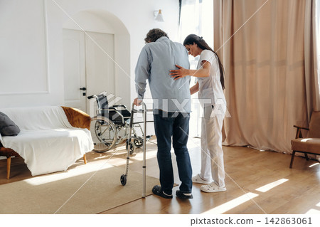 Elderly man using a walker assisted by a caregiver in a bright indoor setting with a wheelchair and comfortable furniture visible in the background Elderly man using a walker assisted by a caregiver in a bright indoor setting with a wheelchair and comfortable furniture visible in the background 142863061