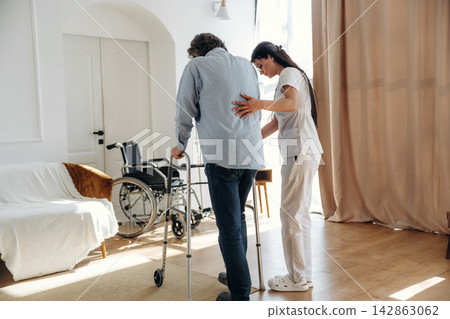 Female caregiver assists elderly man using walker in bright living room with wheelchair and comfortable furniture visible in the background 142863062