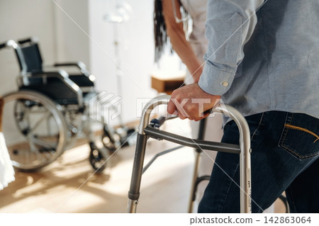 Individual using a walker for support while standing in a bright room with a wheelchair and medical equipment visible in the background Individual using a walker for support while standing in a bright room with a wheelchair and medical equipment visible in the background 142863064