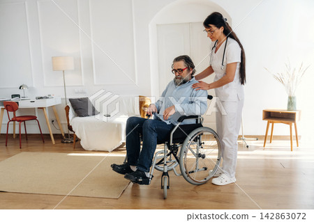 Female caregiver assists elderly man in wheelchair inside a modern living room with wooden flooring, light-colored furniture, and natural light from windows 142863072