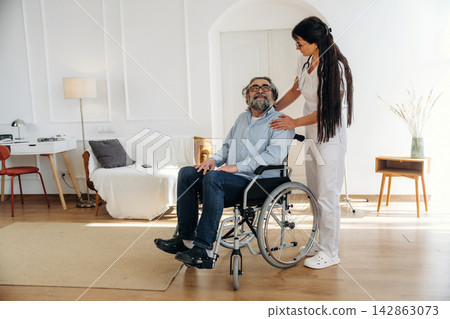 Caregiver assisting elderly man in wheelchair inside a bright living room with modern furniture, featuring a lamp, table, and decorative plants in the background Caregiver assisting elderly man in wheelchair inside a bright living room with modern furniture, featuring a lamp, table, and decorative plants in the background 142863073