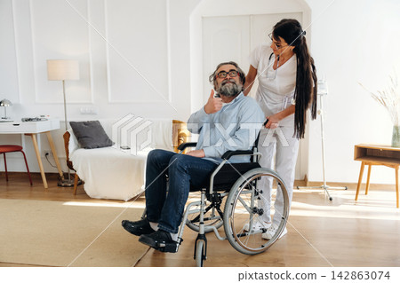 Middle-aged man in wheelchair gives thumbs up while female caregiver assists him in a bright, modern living room with a sofa and decorative elements Middle-aged man in wheelchair gives thumbs up while female caregiver assists him in a bright, modern living room with a sofa and decorative elements 142863074