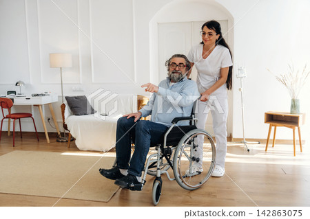 Elderly man in wheelchair being assisted by caregiver in a bright, modern living room with a sofa, lamp, and wooden furniture in the background Elderly man in wheelchair being assisted by caregiver in a bright, modern living room with a sofa, lamp, and wooden furniture in the background 142863075
