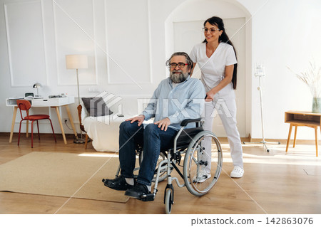 Female caregiver assists elderly man in wheelchair inside a bright, modern living room with wooden flooring and minimalistic furniture Female caregiver assists elderly man in wheelchair inside a bright, modern living room with wooden flooring and minimalistic furniture 142863076