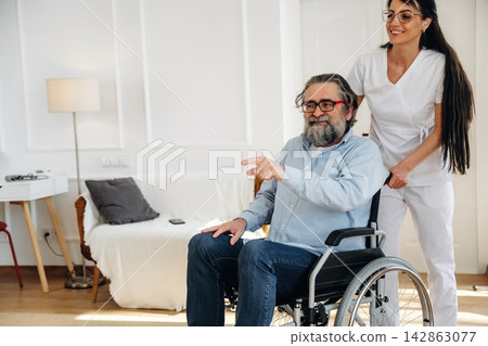 Male patient in wheelchair being assisted by female caregiver in white uniform inside a bright, modern living room with comfortable furniture and soft lighting Male patient in wheelchair being assisted by female caregiver in white uniform inside a bright, modern living room with comfortable furniture and soft lighting 142863077