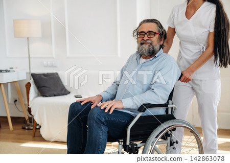 Elderly man in wheelchair smiles while being assisted by caregiver in white uniform inside a bright, modern living room with soft furnishings and wooden flooring 142863078