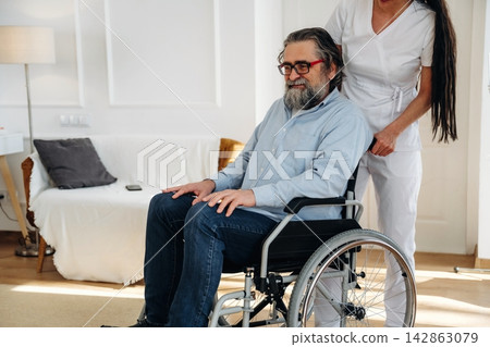 Middle-aged man in wheelchair assisted by caregiver in white uniform inside a bright, modern living room with comfortable furniture and soft lighting 142863079