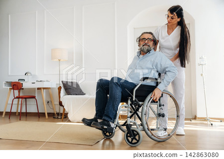 Female caregiver assisting elderly man in wheelchair inside a modern living room with wooden floor, lamp, and furniture in the background Female caregiver assisting elderly man in wheelchair inside a modern living room with wooden floor, lamp, and furniture in the background 142863080